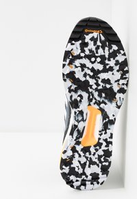 Semelle de chaussure de sport noire et blanche avec un motif texturé et des accents orange, présentant des crampons profonds pour la traction et la stabilité.