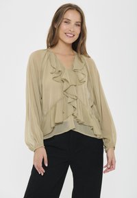 Blusa beige trasparente con un design a strati e volant, maniche lunghe con polsini arricciati e scollatura a V, abbinata a pantaloni neri.