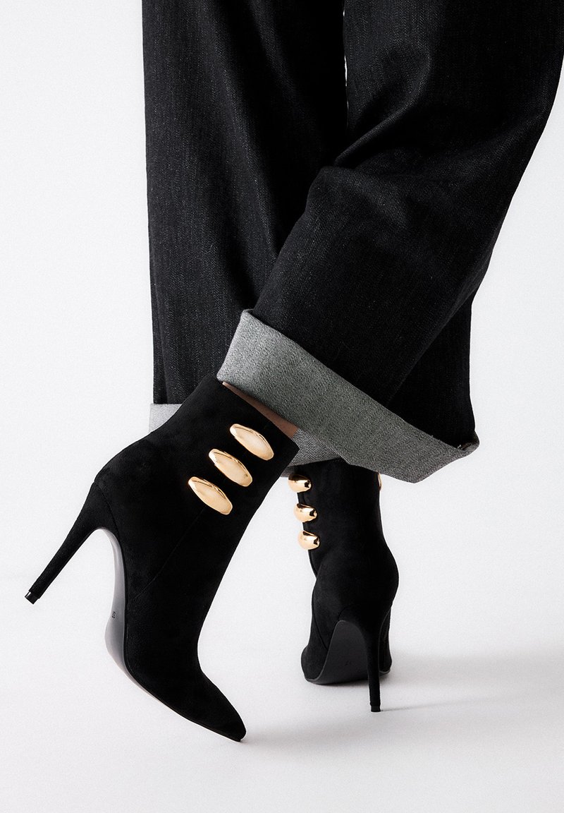 Botines de ante negro de tacón alto y ancho con tres acentos ovalados dorados, puntera afilada y un tacón de aguja estilizado, combinados con pantalones de denim con puños.