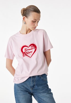 Femme aux cheveux en chignon portant un t-shirt rose clair avec un cœur rouge et le texte « Forever Love », associée à un jean bleu, regardant vers le bas.