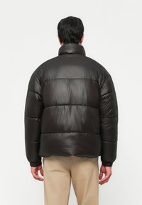 Giacca a vento nera con una superficie liscia e lucida, dotata di un colletto alto e polsini elasticizzati. Design spesso trapuntato e imbottito.
