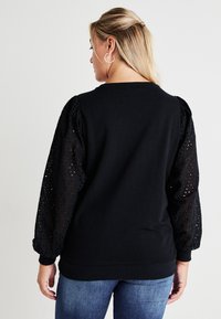Black sweatshirt with structured body and textured lace sleeves. The sleeves feature eyelet patterns, adding contrast to the solid fabric.