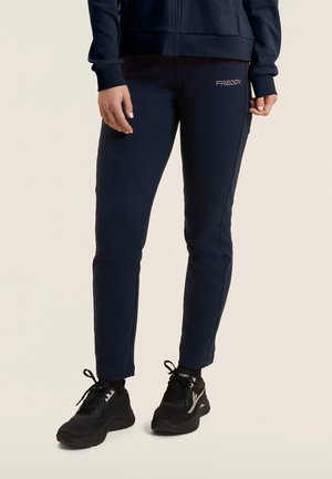 Persona con joggers ajustados azul marino con el logo "FREDDY" en el muslo y zapatillas negras con detalles blancos, de pie sobre un fondo claro.