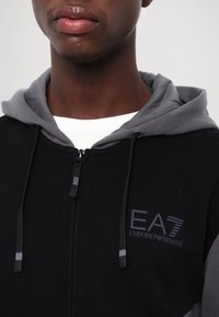 Svart zip-up hoodie med gråa detaljer. Har en dragsko på huva, mjukt material och EA7 Emporio Armani-logotyp på bröstet.