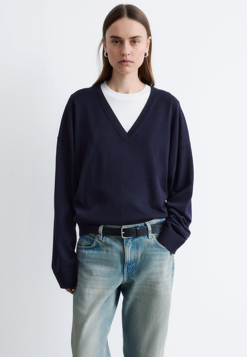 Ung kvinde iført marineblå V-hals trøje over hvid skjorte, stoppet ned i lyseblå jeans med sort bælte, stående foran en ensfarvet baggrund.