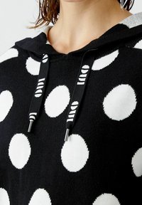 Hoodie noir avec de grands pois blancs, doté d'un cordon de serrage avec des accents argentés et une texture douce. Détails de couture visibles au niveau du col.