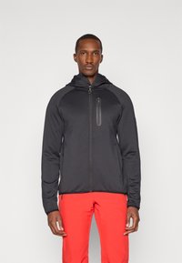 Melnā zipa jakā ar kapuci, ar faktūras apdari, kontrastējošām plecu daļām un mazu krūšu kabatu. Sniegta kopā ar sarkaniem biksēm.