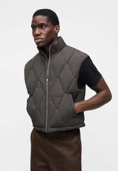 Colete cinzento acolchoado com gola alta e fecho de correr frontal. Apresenta costura em forma de diamante e design sem mangas, usado por cima de uma camisa preta.
