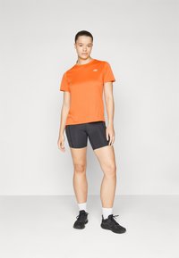 Oranžna športna majica s kratkimi rokavi, majhen logotip na prsi. Vse skupaj je ujemano s črnimi oprijetimi kratkimi hlačami in črnimi supergami z belimi nogavicami.