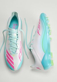 Λευκά και τιρκουάζ ποδοσφαιρικά παπούτσια με ροζ γράμματα "FSO", κορδόνια λυμένα, που δείχνουν την υφή της σόλας και τις ρίγες στην πλευρά.