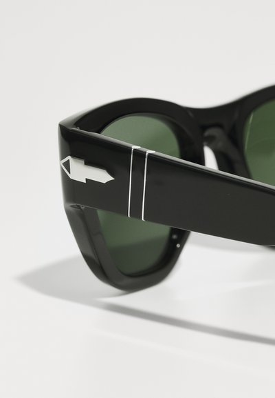 Čierne slnečné okuliare so zelenými sklá. Majú lesklý povrch, nadrozmerný obdĺžnikový tvar a charakteristický dizajn šípu na ramene.