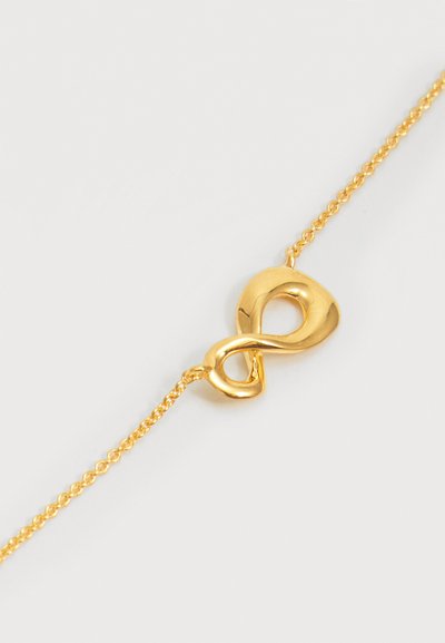 Βραχιόλι αλυσίδα από χρυσό με ένα απαλό, γυαλιστερό μενταγιόν με το σύμβολο του άπειρου σε ανοιχτό φόντο.