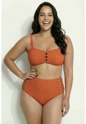 Glunderende vrouw met lang donker haar draagt een gestructureerde oranje bikini met hoge taille en uitgesneden details, poserend tegen een effen lichte achtergrond.