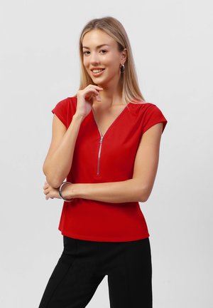 Rotes Kurzarmoberteil mit einem Reißverschluss-V-Ausschnitt, aus glattem Stoff und mit taillierter Silhouette, kombiniert mit schwarzen Hosen.