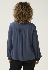 Blouse à manches longues bleu marine avec une coupe décontractée, des poignets élastiques et des détails froncés au dos. Tissu lisse avec une finition mate.