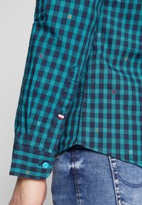 Camicia a quadri turchese e blu scuro con accenti multicolori, polsino abbottonato e dettagli di cuciture visibili. Progettata per un uso casual.
