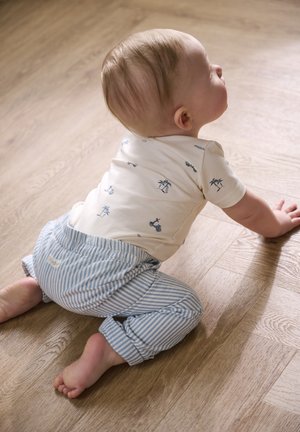 Baby met licht haar, gekleed in een wit shirt met blauwe prints en blauw gestreepte broek, die kruipt over een houten vloer.