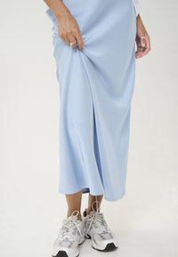 Hellblaues Midikleid aus glattem Stoff mit seitlichem Schlitz, kombiniert mit weißen und silbernen Sportschuhen. Die Hände greifen leicht den Stoff.