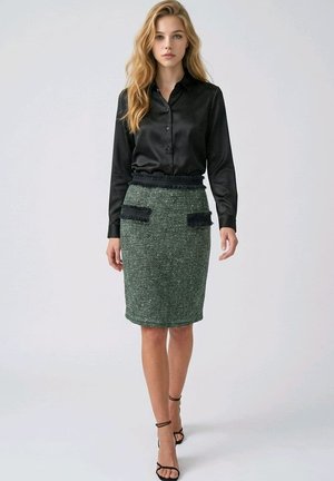 Mujer con camisa negra de satén abotonada, falda verde texturizada hasta la rodilla con bolsillos negros con flecos, y tacones negros con tiras, de pie contra un fondo liso.