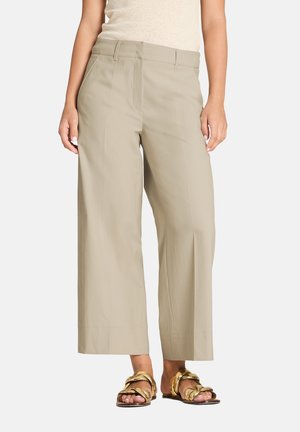 Vrouw die beige cropped wijde broekspijpen en gouden gevlochten platte sandalen draagt, staand tegen een effen witte achtergrond.