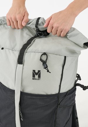 Mains sécurisant la boucle supérieure d'un sac à dos roller Millet gris et noir avec poches latérales en filet sur fond blanc.