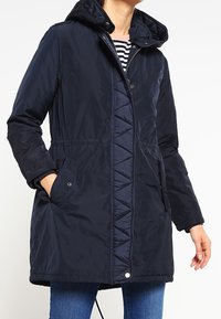 Námětčná modrá prošívaná bunda s kapucí, která má přední zip, boční kapsy a trojúhelníkový design podél zapínání na knoflíky.