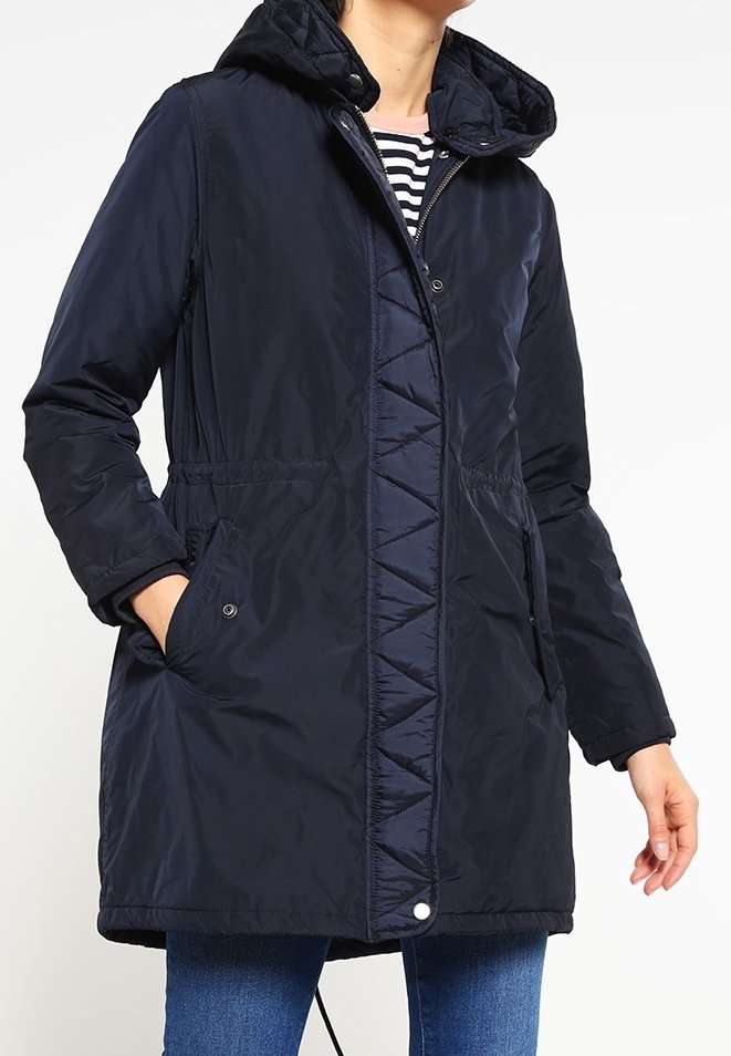 Námětčná modrá prošívaná bunda s kapucí, která má přední zip, boční kapsy a trojúhelníkový design podél zapínání na knoflíky.