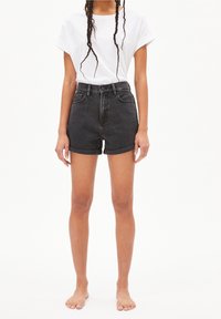 Kvinna med långt flätat hår, klädd i en vit t-shirt och höga svarta jeansshorts, står barfota mot en vit bakgrund.