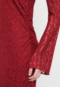 Κόκκινο φόρεμα από δαντέλα με μακριά μανίκια σε σχήμα καμπάνας, που δείχνει λεπτομέρεια με πιέτες στη μέση και ένα χέρι που ακουμπά απαλά στο πλάι.