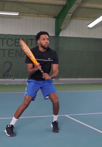 Τενίστρια σε μαύρο μπλουζάκι και μπλε σορτς κρατάει μια πορτοκαλί ρακέτα, έτοιμη να παίξει σε κλειστό γήπεδο με πράσινα δίχτυα και μπλε δάπεδο.