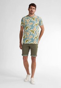 Vaaleansininen lyhythihainen paita, jossa on kukka- ja lehtikuvioita, yhdistetty oliivinvihreisiin shortseihin. Asun täydentävät valkoiset lenkkarit.