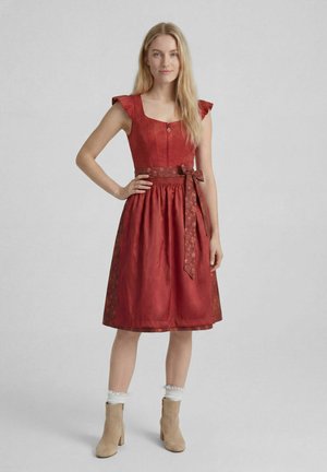 Frau mit langen blonden Haaren, die ein rotes knielanges Kleid mit gebundenem Gürtel und beigen Stiefeletten trägt, steht vor einem schlichten Hintergrund.