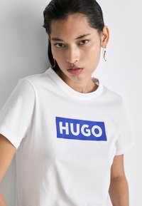 Bílý bavlněný tričko s modrým obdélníkovým logo, na kterém je tučný bílý nápis "HUGO". Klasický design s kulatým výstřihem.