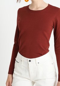 Rostfarbener Langarm-Oberteil mit Rundhalsausschnitt, aus geripptem Stoff gefertigt, kombiniert mit weißen hochgeschnittenen Jeans, die über vordere Taschen verfügen.