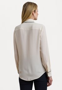 Light beige blouse made of sheer fabric with a button-down design, long sleeves, and contrasting black piping on cuffs. Curved hem.