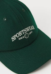 Groene wolcaps met geborduurde tekst "SPORTSWEAR" en een grafisch tennisbal. Heeft een gebogen rand en gestikte details langs de rand.