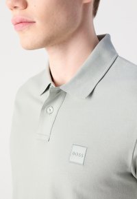 Nærbilde av en person som har på seg en lys grå polo skjorte med krage, knapp og en liten "BOSS" logo på brystet.