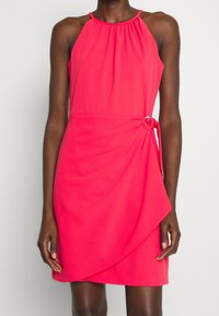 Robe sans manches fuchsia avec un col froncé et un détail de nœud sur le côté, présentant un design de portefeuille asymétrique et une texture lisse.