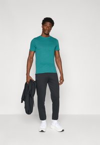 Türkiisel lühikeste varrukatega T-särk, slim-fit mustad joggerid elastsete mansettidega ja valged spordijalatsid. Tumeda jope on visatud tema käele.
