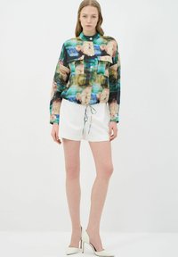 Camisa de seda con botones, que presenta un estampado floral abstracto en azul, verde y melocotón, con dos bolsillos frontales y un dobladillo ajustable. Pantalones cortos blancos.