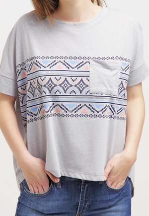 Vrouw draagt een lichtgrijs shirt met korte mouwen met geometrische patronen, een borstzak en een blauwe spijkerbroek, handen in de voorzakken gestopt.