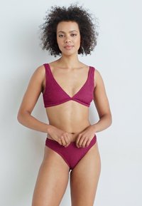 Bikinis komplektas tamsiai bordo spalvos, pagamintas iš tekstūruotos, banguotos medžiagos. Viršutinė dalis turi plačius petnešas ir trikampį iškirpimą, derinama su aukštu juosmeniu.