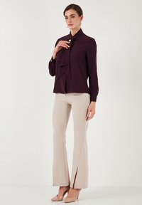 Blusa viola scuro con un davanti arricciato, maniche lunghe e chiusure a bottoni, abbinata a pantaloni beige a zampa d'elefante con spacco laterale.