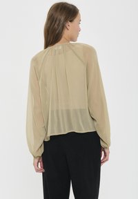 Blusa trasparente a maniche lunghe di un leggero beige con dettaglio arricciato sul retro. Vestibilità ampia, tessuto testurizzato e scollo rotondo. Abbinata a pantaloni scuri.