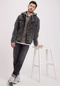 Czarna kurtka dżinsowa na szarej bluzie z kapturem, w zestawieniu z ciemnymi dżinsami. Model opiera się o białe stołek, mając na sobie szare trampki.