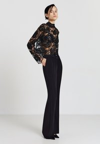 Schwarzes Spitzenoberteil mit langen Ärmeln und floralem Stickmuster, kombiniert mit hoch taillierter schwarzer Hose. Die Hose hat einen geraden Schnitt und eine leichte Ausweitung am Saum.