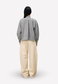 Frau mit langen schwarzen Haaren, die eine kariert gemusterte Bluse mit langen Ärmeln und hochtaillierte, weite, beigefarbene Hosen trägt, mit dem Rücken zur Kamera vor einem einfarbigen Hintergrund.