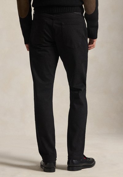 Muž nosiaci čierne slim fit nohavice, čierne topánky a čierny sveter s hnedými nášivkami na lakťoch, stojaci na neutrálnom pozadí.