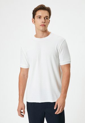 Camiseta de manga corta blanca con un patrón texturizado de diamantes, cuello redondo y ribete de contraste en las mangas. Combinada con pantalones oscuros.
