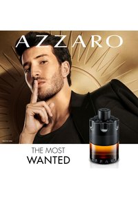 Man met donker haar en stoppels die een vinger op zijn lippen houdt, zwarte jas, met een amberkleurige en zwarte Azzaro geurflacon naast hem.